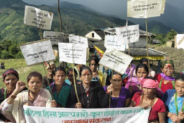 A group of Nepali women hold a banner and placards as they protest in front of their remote village, with hills in the background