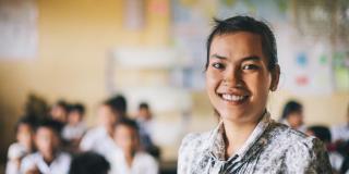 Primary school teacher Souy Kran smiles as she stands at the front of her classroom; behind her, rows of schoolchildren work at their desks.