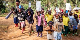 A group of young children carry teaching materials as they walk outside, led by their teacher