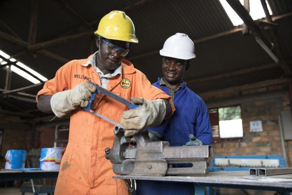 A student in overalls and safety gear operates machinery in a metal workshop, overseen by his instructor