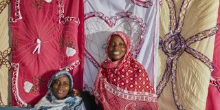 Two female students pose in front of a wall of hanging fabrics at the Subra Tailoring Enterprise