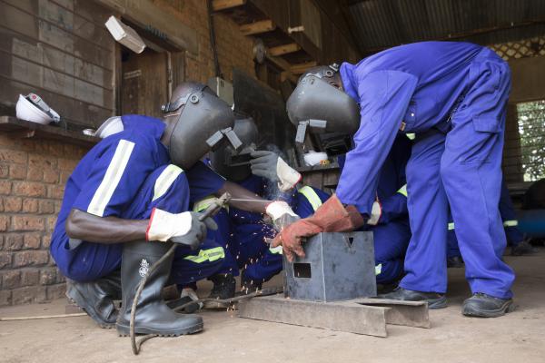 A group of trainee welders in full protective equipment help each other to practise welding a metal box
